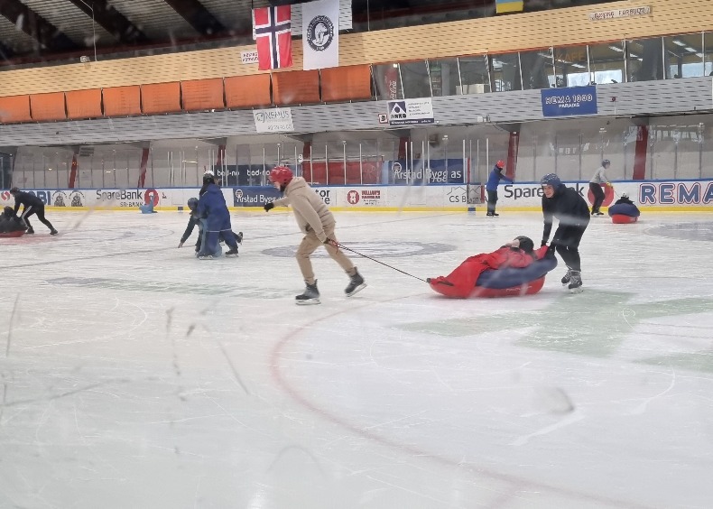En person sitter i en pulk og blir trukket av to personer rundt på isen